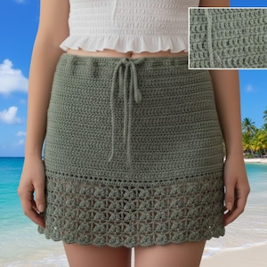 May include: A sage green crochet skirt with a drawstring waist and a scalloped hem. The skirt is handmade with a detailed openwork pattern at the bottom. The skirt is shown against a beach background.