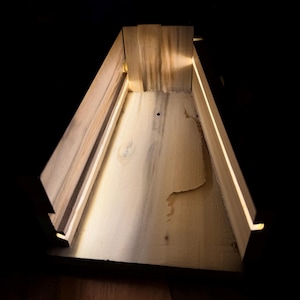 May include: A wooden, triangular-shaped box with integrated LED lighting. The box is constructed from light-colored wood, with visible grain and a natural finish. The interior is illuminated by a warm, soft glow from the embedded lights, creating a modern, minimalist aesthetic.