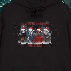 Könnte beinhalten: Schwarzer Kapuzenpullover mit einem Aufdruck von Horrorfilmfiguren mit Weihnachtsmützen und Waffen. Der Text "Slashing Through The Snow" befindet sich über den Figuren. Schneeflocken sind im Design enthalten.