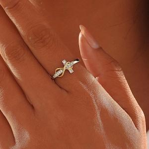 May include: A silver and gold ring featuring a cross design with clear gemstones. The ring has an infinity symbol incorporated into the design. The ring is on a person's finger.