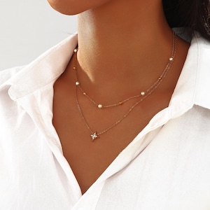 May include: A layered gold-toned necklace with a delicate design. The top layer features small pearls and gold beads, while the lower layer showcases a star-shaped pendant with clear crystals. The necklace is worn over a white collared shirt.