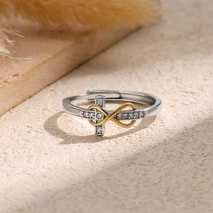 May include: A silver-tone ring with a gold-tone infinity symbol and a cross design. The cross and infinity symbol are embellished with small, clear gemstones. The ring is set against a neutral background.