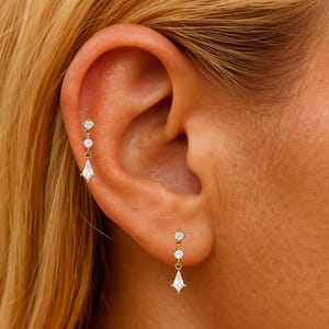 May include: Gold-toned earrings with a dangling design. Each earring features three clear, sparkling stones, with a larger, diamond-shaped stone at the bottom. The earrings are worn on an ear, showcasing their delicate and elegant style.