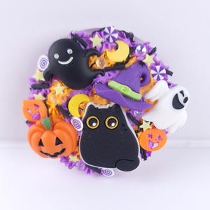 Könnte beinhalten: Ein kreisförmiges Halloween-Dekorationselement mit einer schwarzen Katze, Kürbissen, Geistern und einem Hexenhut. Das Design verwendet lila, orange, gelbe und weiße Farben und erzeugt eine festliche Atmosphäre. Wahrscheinlich eine Brosche oder ein dekoratives Ornament.
