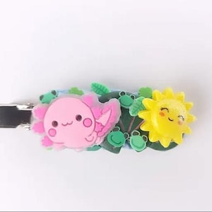 May include: A decorative hair clip featuring a pink axolotl, green leaves, and a yellow smiling sun. The clip is made of resin and has a silver metal clasp. The axolotl has a cute expression, and the sun has a smiling face.