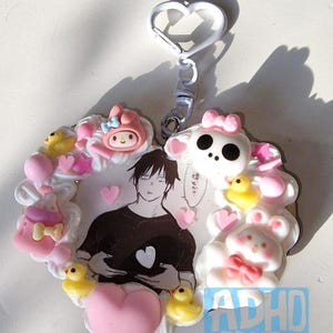 Könnte beinhalten: Ein herzförmiger Schlüsselanhänger mit einer Cartoon-Illustration einer Person und verschiedenen dekorativen Elementen. Das Design umfasst eine My Melody-Figur, einen Totenkopf, Enten, Herzen und den Text "ADHD no Jutsu".