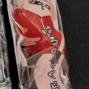 May include: A plush doll in a clear plastic package. The doll has a pink head, red clothing, and black text. The doll's design includes a face with eyes and a mouth. The doll is in a vertical position.