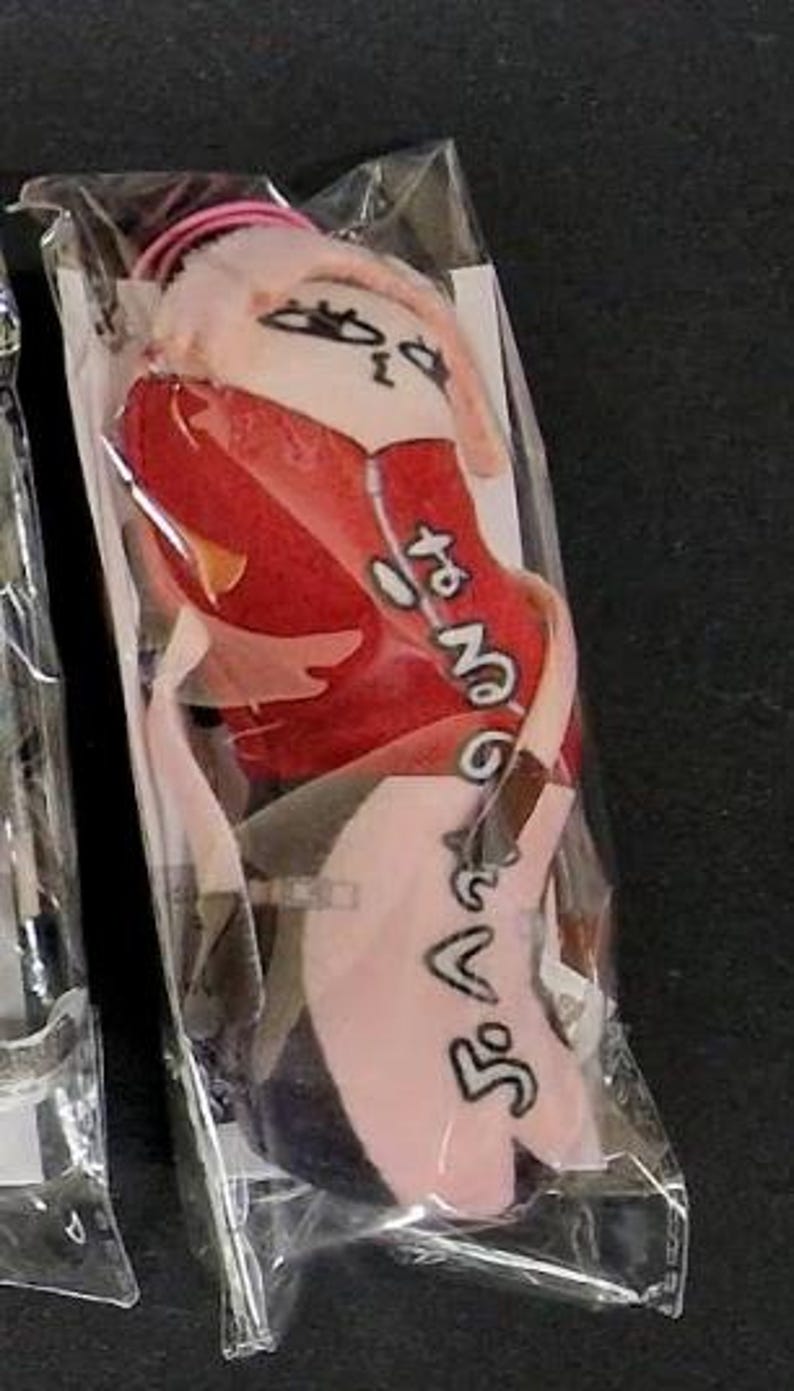 May include: A plush doll in a clear plastic package. The doll has a pink head, red clothing, and black text. The doll's design includes a face with eyes and a mouth. The doll is in a vertical position.