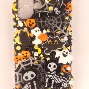 May include: A black phone case decorated with Halloween-themed embellishments. The case features a spiderweb design, pumpkins, ghosts, bats, and skeletons. The decorations are in orange, white, and black, with glitter accents.