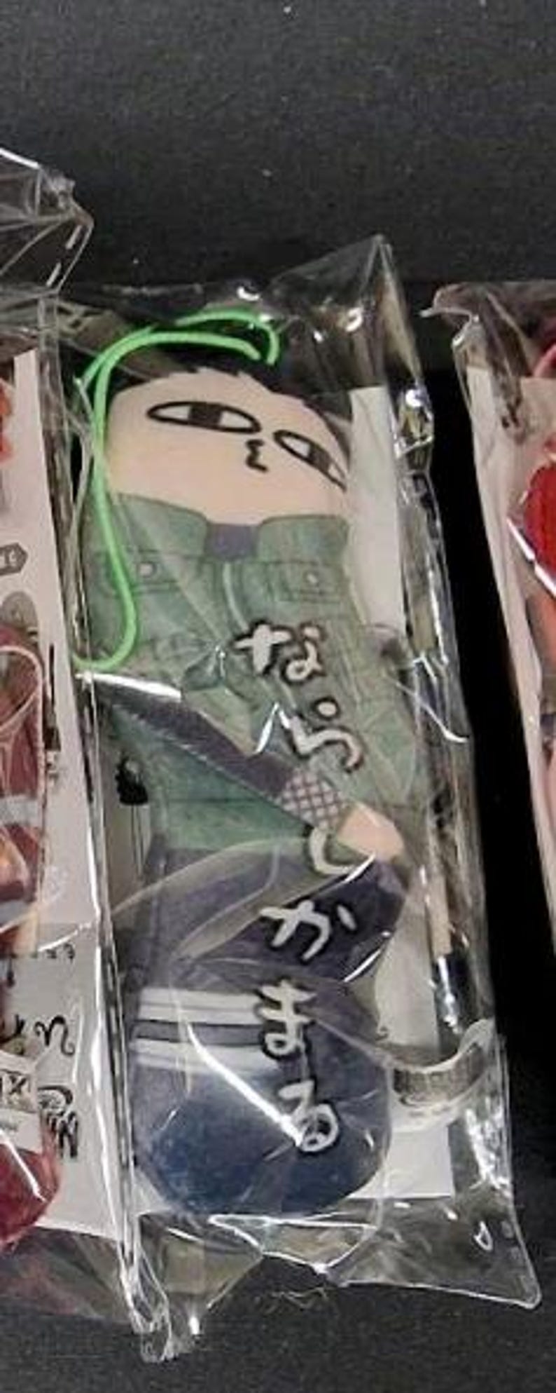 May include: A plush keychain in a clear plastic package. The plush depicts a cartoon character wearing a green jacket and blue pants. Japanese text is printed on the plush. A green string is attached to the top of the plush.