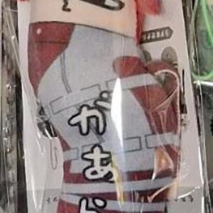 May include: A plush doll of a character with red hair and a distinctive face, packaged in clear plastic. The doll is primarily red, white, and gray, with Japanese text on the front. A small white tag with the word "MAX" is attached.