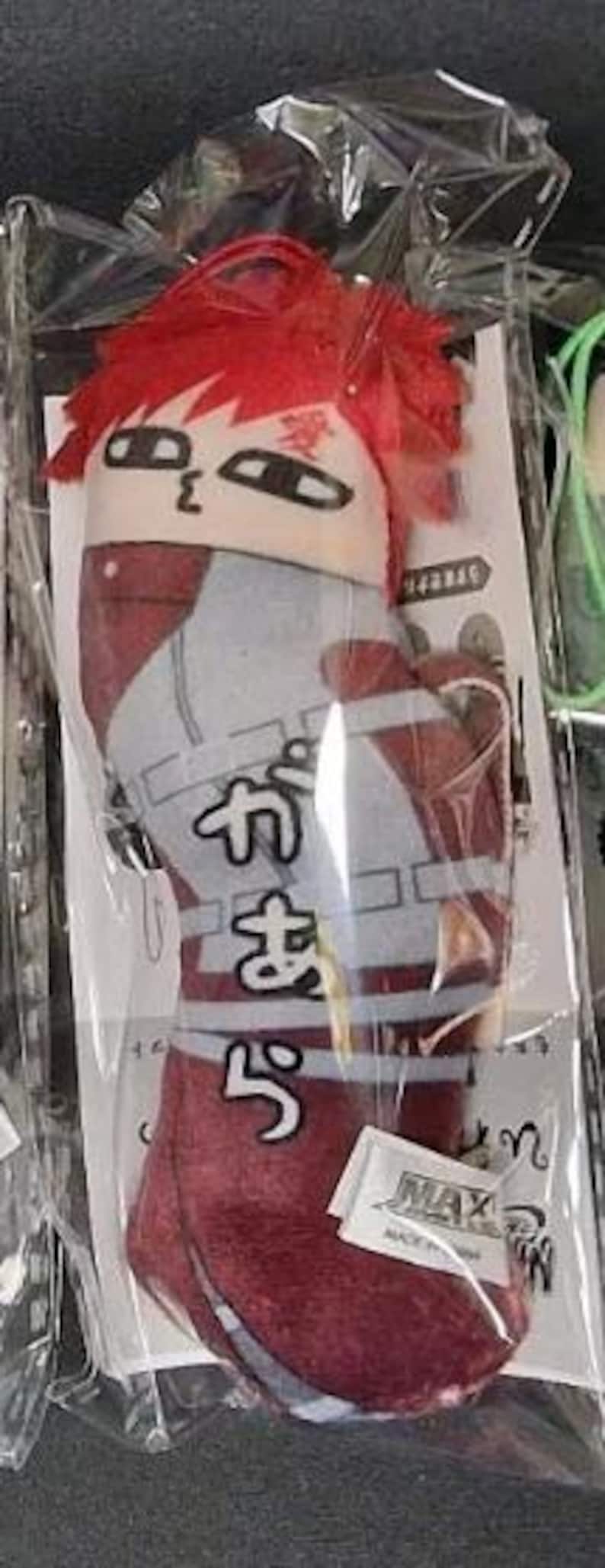 May include: A plush doll of a character with red hair and a distinctive face, packaged in clear plastic. The doll is primarily red, white, and gray, with Japanese text on the front. A small white tag with the word "MAX" is attached.