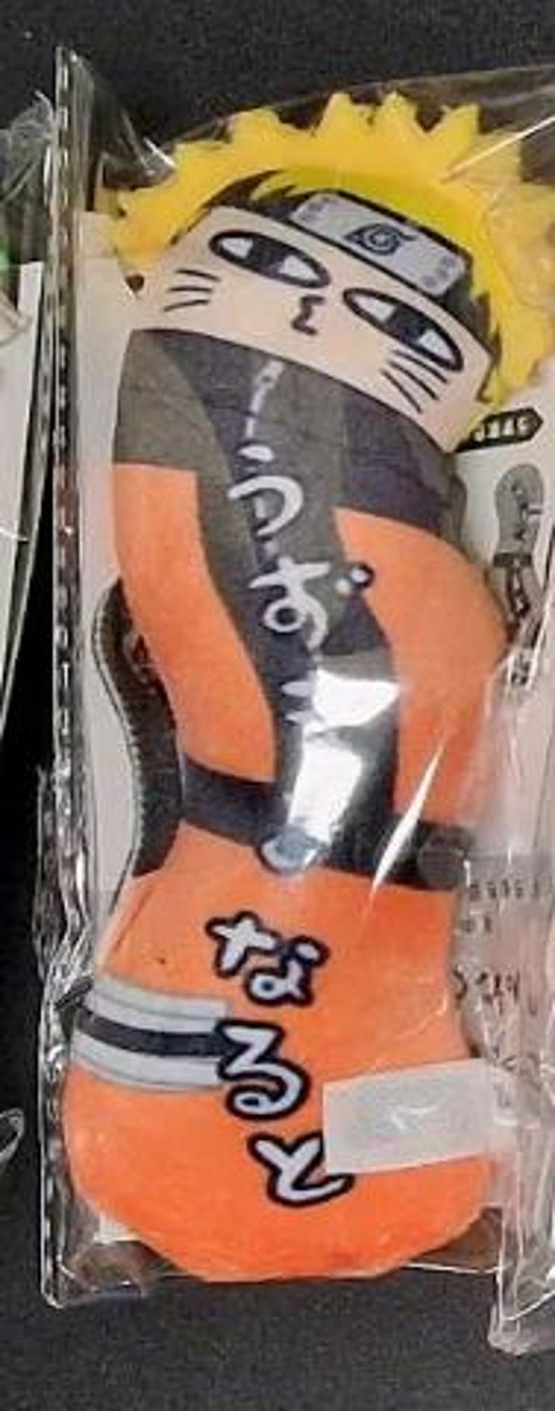 May include: Orange and black plush toy in a clear plastic package. The toy is shaped like a character with yellow hair, a headband, and Japanese text. The character is wearing an orange jumpsuit with black accents and a white and blue belt.