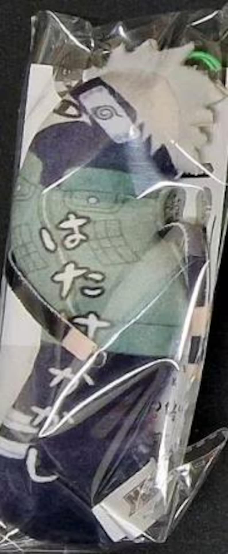 May include: A plush toy of a character from an anime series, packaged in clear plastic. The toy features a white-haired character with a headband and a green and blue outfit. The Japanese text "はたけ" is visible on the outfit.