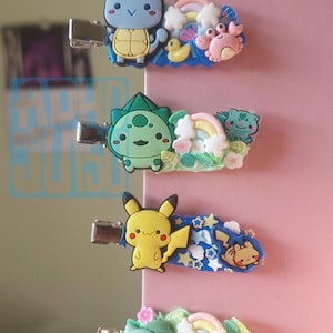 May include: Four colorful hair clips featuring cartoon characters. The clips include a blue turtle, a green Bulbasaur, a yellow Pikachu, and a light green cat. Each clip has a unique design with rainbows, flowers, and other decorative elements.