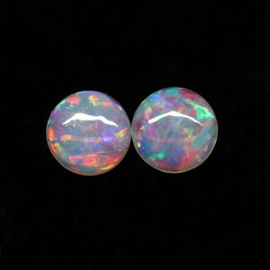 Puede incluir: Dos ópalos redondos con un vibrante juego de colores, mostrando destellos de rojo, verde y azul. Los ópalos están sobre un fondo negro, resaltando sus cualidades iridiscentes. Estas piedras preciosas son ideales para la creación de joyas.