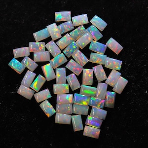 Puede incluir: Una colección de ópalos rectangulares con destellos de colores iridiscentes. Los ópalos son de color blanco lechoso y están dispersos sobre una superficie negra. Los ópalos tienen una superficie pulida y lisa y son de varios tamaños.