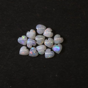 Puede incluir: Un conjunto de ópalos en forma de corazón con destellos iridiscentes de color. Los ópalos son blancos con un juego de colores, incluyendo toques de azul, verde y rosa. Las piedras preciosas están dispuestas sobre una superficie negra.