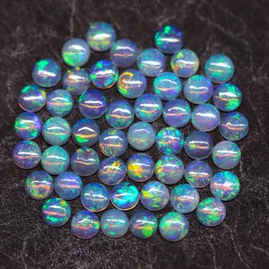Könnte beinhalten: Eine Ansammlung runder Opale mit irisierenden Farben von Blau, Grün und Lila. Die Opale sind auf einer dunklen Oberfläche verstreut und zeigen ihr lebendiges Farbspiel. Diese Edelsteine eignen sich ideal für die Schmuckherstellung.