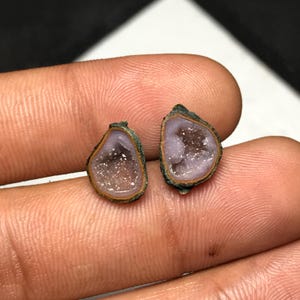 May include: Two small, irregular, teardrop-shaped geode slices featuring a light purple, crystalline center surrounded by a thin, dark brown outer rim. The stones have a rough, natural texture and a sparkling, glittery appearance within the hollowed-out centers.