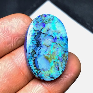 May include: An oval-shaped boulder opal gemstone featuring a vibrant play of color with dominant shades of electric blue, turquoise, and deep violet. The surface displays a complex network of dark, branching matrix veins throughout the stone.