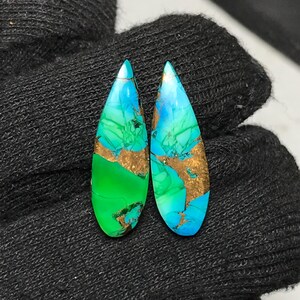 May include: Two teardrop-shaped cabochons featuring a vibrant blend of bright blue and green hues with prominent metallic copper-colored veining throughout the polished surfaces, showcasing a smooth, glossy finish and distinct color transitions.