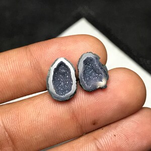 Könnte beinhalten: Zwei kleine, unregelmäßige, tropfenförmige Geodenscheiben mit dunkelvioletten, kristallinen Innenräumen und einer funkelnden, glitzerartigen Textur. Die Außenkanten dieser Steine sind durch ein glattes, hellgraues und weißes Mineralband definiert.