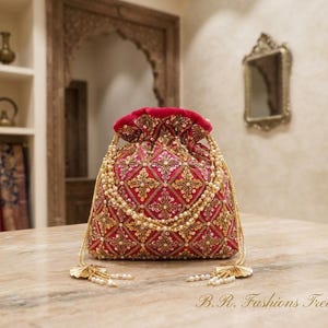 Könnte beinhalten: Ein fuchsiafarbener Beutel mit Kordelzug, verziert mit einer rautenförmigen Stickerei aus Goldfaden, Pailletten und kleinen weißen Perlen. Die Tasche hat einen Perlenhenkel und zwei goldfarbene Quasten mit Perlen an den Kordelenden.