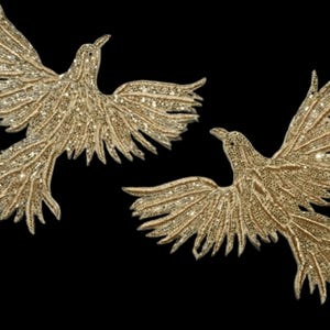 May include: Two gold-coloured bird-shaped appliques with outstretched wings, embellished with sparkling details. The birds are set against a black background, creating a striking contrast. The appliques are likely for decorative purposes.