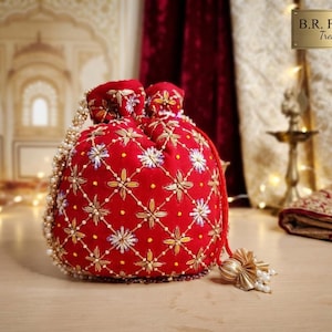 Peut inclure: Une pochette à cordon en velours rouge ornée d'un motif en treillis brodé doré et blanc avec des accents floraux. Le sac comprend une anse en perles et une fermeture à pompon doré. Le texte B.R. Fashions Treasures est visible.