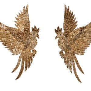 May include: Two gold-coloured bird-shaped embellishments with outstretched wings. The birds have detailed feather patterns and are adorned with small beads. The embellishments are likely for decorative purposes, such as on clothing or accessories.