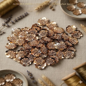 Peut inclure: Une collection d'ornements dorés en forme de fleurs, dispersés sur une surface en lin neutre. Les ornements ont un design détaillé et superposé. L'image comprend le texte "AURORA EMBELLISHMENTS - Handcrafted Series."