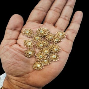 May include: A collection of gold-colored, flower-shaped embellishments, each with a central clear gemstone. The filigree design of the flowers is visible, and they are displayed against a dark background.