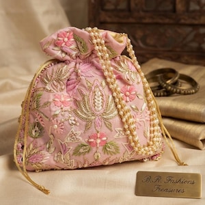 Könnte beinhalten: Ein hellrosa Beutel mit Kordelzug und aufwendiger Blumenstickerei in Rosa-, Grün- und Goldtönen. Die Tasche hat einen Perlenhenkel und goldene Kordeln. Ein kleines rechteckiges goldenes Schild trägt die Aufschrift B.R. Fashions Treasures.