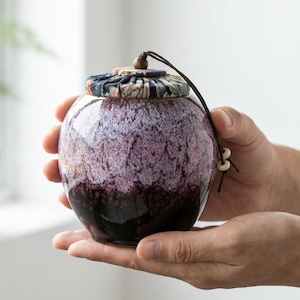 Puede incluir: Un pequeño tarro de cerámica redondo con tapa. El tarro tiene un degradado de colores, que va del morado oscuro en la base a un morado más claro y moteado en la parte superior. La tapa tiene un diseño floral. Una cuerda negra con una cuenta de madera está adjunta.