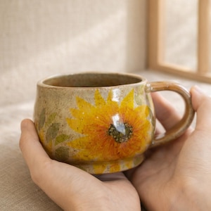 May include: A ceramic mug with a sunflower design. The mug is beige with a large yellow and orange sunflower painted on the front. Green leaves are painted around the sunflower. The mug has a rounded shape and a brown handle.