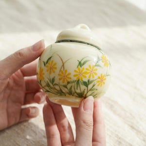 May include: A small, cream-coloured ceramic jar with a lid, hand-painted with yellow daisy-like flowers and green leaves. The jar is held by two hands, showcasing the delicate floral design. The lid has a small handle.
