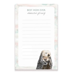 May include: A white notepad with a watercolor background of pink and blue. The notepad has a lined section for writing and the text "BEST MOM EVER" and "amanda young" at the top. The bottom of the notepad features a watercolor illustration of two sea otters hugging.