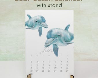 May include: A 2027 Ocean Calendar with stand featuring a watercolor illustration of two blue dolphins. The page displays the month of June with a grid of dates from 1 to 30. The text 2027 Ocean Calendar with stand is printed at the top.