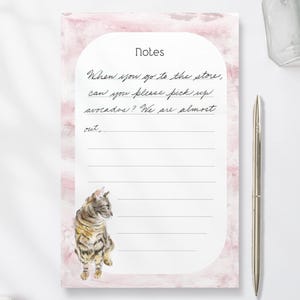 Peut inclure: Un bloc-notes avec un motif marbré rose et le mot "Notes" en haut. Le bloc-notes comporte des lignes pour écrire et une illustration à l'aquarelle d'un chat. Le texte sur le bloc-notes se lit comme suit : "Quand tu vas au magasin, peux-tu acheter des avocats ? On n'en a presque plus."