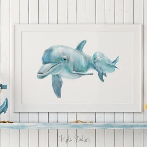 Pu&ograve; includere: Una stampa ad acquerello incorniciata con due delfini azzurri che nuotano insieme. L'opera d'arte &egrave; esposta su una mensola bianca con decorazioni nautiche, tra cui un'ancora blu e un gabbiano decorativo. Il testo "Triple Studio" &egrave; visibile.
