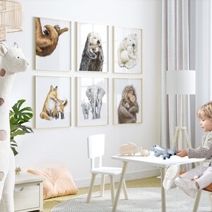May include: A collection of six framed animal art prints, featuring watercolor illustrations of a sloth, sea otter, polar bear, fox, elephant, and monkey with their young. The prints are displayed on a white wall in a child's room.