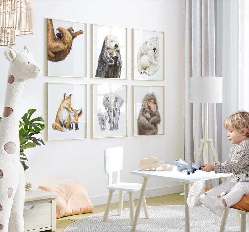 May include: A collection of six framed animal art prints, featuring watercolor illustrations of a sloth, sea otter, polar bear, fox, elephant, and monkey with their young. The prints are displayed on a white wall in a child's room.
