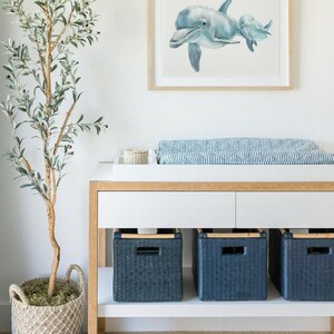 Pu&ograve; includere: Una stampa ad acquerello incorniciata di due delfini in tonalit&agrave; di blu &egrave; appesa sopra un fasciatoio bianco. Il tavolo ha tre cesti portaoggetti in vimini blu navy. Un albero di ulivo in vaso in un cesto di vimini &egrave; a sinistra.