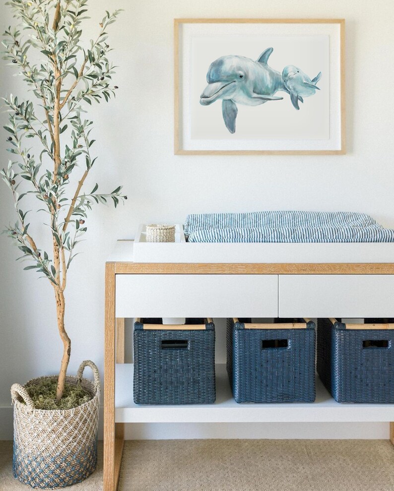 Pu&ograve; includere: Una stampa ad acquerello incorniciata di due delfini in tonalit&agrave; di blu &egrave; appesa sopra un fasciatoio bianco. Il tavolo ha tre cesti portaoggetti in vimini blu navy. Un albero di ulivo in vaso in un cesto di vimini &egrave; a sinistra.