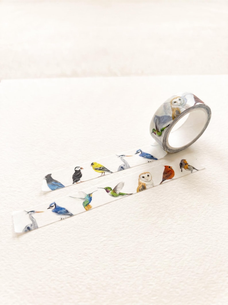 May include: A roll of white washi tape featuring a repeating pattern of various colourful birds, including a blue jay, puffin, goldfinch, heron, hummingbird, barn owl, cardinal, and robin, printed in a detailed, realistic watercolour style.