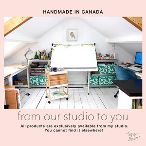 Pu&ograve; includere: Luminoso studio d'arte con tavolo da disegno, carrello e scaffali. Il testo "HANDMADE IN CANADA" e "from our studio to you" sono visibili. Lo studio &egrave; pieno di materiali artistici e un computer.