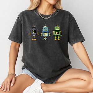 May include: Dark gray t-shirt featuring three colorful robot illustrations. Each robot has a different number displayed on its chest: 01, 10, and 11. The robots are in purple, blue, and green with yellow accents.