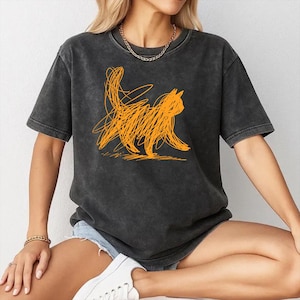 Puede incluir: Camiseta gris oscuro con un diseño de gato garabateado en naranja. La camiseta de manga corta tiene un corte holgado y un aspecto ligeramente descolorido. El gráfico del gato es una ilustración simple y abstracta.