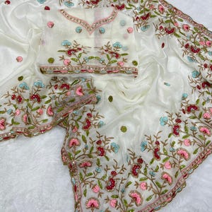 Peut inclure: Un sari blanc avec des broderies florales complexes dans les tons de rose, de rouge et de bleu. Le sari comprend un chemisier assorti avec des détails similaires. Les bords sont ornés d'une bordure délicate et de paillettes, créant un look élégant et festif.